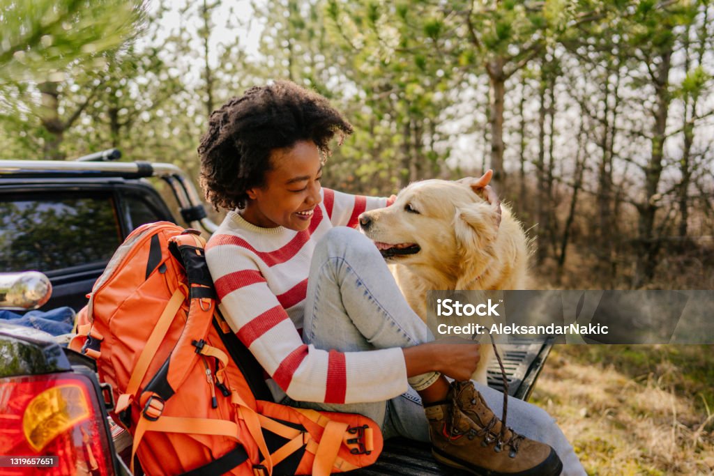 Woman sitting on the back of a vehicle with hiking gear, smiling at her dog in a wooded setting.