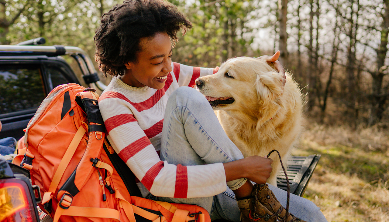 Woman sitting on the back of a vehicle with hiking gear, smiling at her dog in a wooded setting.